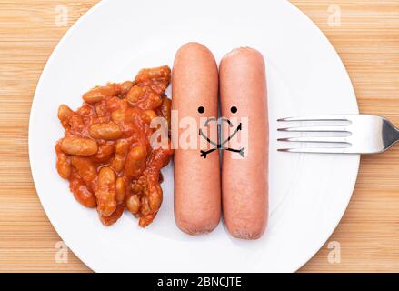Due salsicce grasse con volti tristi, adagiate su un piatto bianco con forchetta e fagioli, immagine concettuale sulla dieta, alimentazione sana e obesità Foto Stock