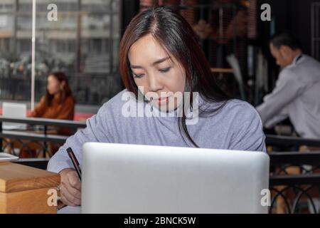 Smart bella donna asiatica intelligente scrivere e annotare le informazioni di sintesi positivo studio e business script saggio di fronte al computer portatile a. Foto Stock