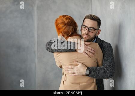 Giovane uomo in occhiali abbracciando la donna e supportandola mentre si levano in piedi contro la parete grigia Foto Stock