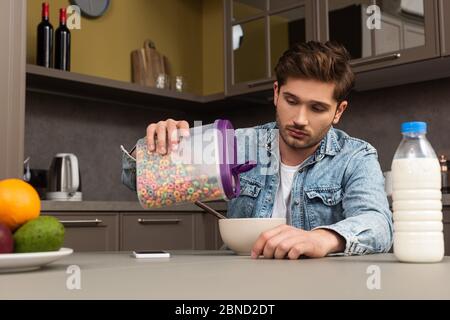Fuoco selettivo dell'uomo che versa i cereali nel recipiente vicino alla bottiglia del latte e della frutta sul tavolo della cucina Foto Stock