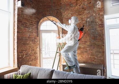 Coronavirus Pandemico. Un disinfettante in tuta protettiva e maschera spruzza disinfettanti in casa o in ufficio. Protezione contro la malattia COVID-19. Prevenzione del virus della polmonite da spandimento con superfici. Foto Stock