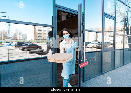 Il proprietario del caffè in maschera medica vicino alla porta con la scheda con la scritta di quarantena che mostra le scatole e guardando la macchina fotografica sulla strada Foto Stock