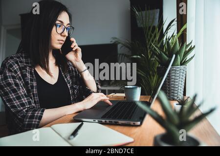 Giovane donna che parla al telefono mentre lavora al laptop. Donna d'affari in conversazione al telefono in ufficio. Concetto di lavoro da casa Foto Stock