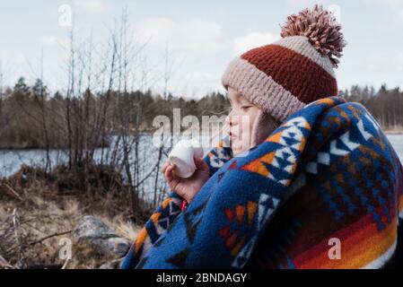 ragazza giovane avvolta in una coperta che mangia un marshmallow mentre campeggio Foto Stock