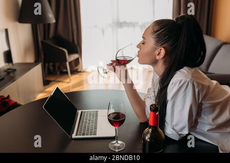 vista laterale della giovane donna che beve vino rosso vicino al computer portatile con schermo vuoto Foto Stock