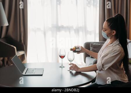 vista laterale di una giovane donna in maschera protettiva che versa il vino in vetro vicino a un computer portatile con schermo vuoto Foto Stock