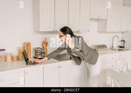 donna d'affari in blazer sopra i pigiami che parlano sullo smartphone e usando il laptop in cucina Foto Stock