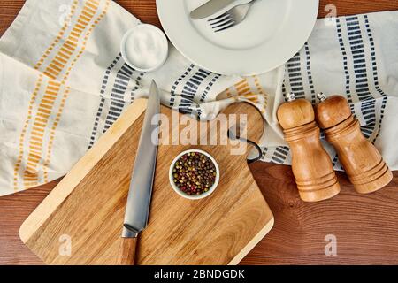 Vista dall'alto del piatto con posate vicino al panno, tagliere, ciotole, pepe e mulini di sale su sfondo di legno Foto Stock