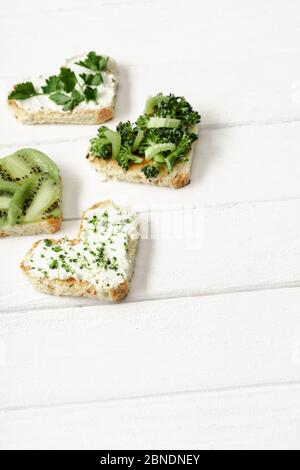 canape a forma di cuore con formaggio cremoso, broccoli, micrograno, prezzemolo e kiwi su superficie di legno bianco Foto Stock