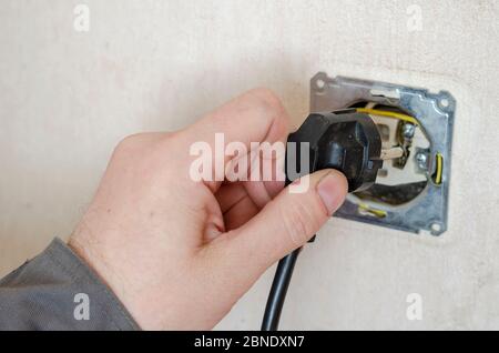 Un elettricista controlla una presa smontata. La mano di un uomo in indumenti da lavoro protettivi porta la spina elettrica alla presa smontata. Pericolo Foto Stock