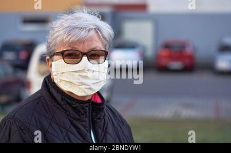 Anziana donna anziana che indossa la casa fatto tessuto bocca viso naso virus maschera fuori, auto sfocate e sfondo edificio. Può essere usato durante il coronavirus Foto Stock