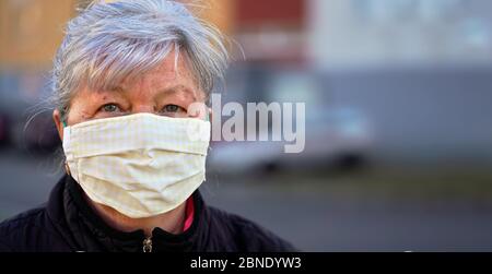 Anziana donna anziana che indossa casa fatto tessuto bocca viso naso virus maschera fuori, sfondo edificio sfocato. Può essere usato durante il coronavirus covid-19 Foto Stock