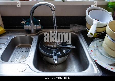 L'acqua corrente di un rubinetto riempie le pentole che si immergerono in un lavello da cucina. I piatti sono impilati e pronti per essere lavati. Foto Stock