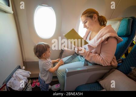 bella madre si siede su una sedia dell'aeroplano e legge un libro. carino toddler sta in piedi davanti a lei. primo piano, messa a fuoco morbida, vista dall'alto. copyspace Foto Stock