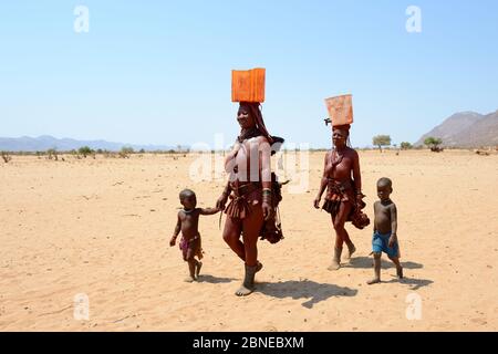 Donne e bambini di Himba che ritornano dal punto d'acqua, trasportando lattine di plastica piene d'acqua, durante la stagione secca, la valle di Marienfuss, il deserto di Kaokoland, Foto Stock