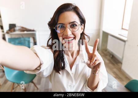 Attraente giovane bruna donna d'affari in piedi nel caffè al coperto, prendendo un selfie Foto Stock