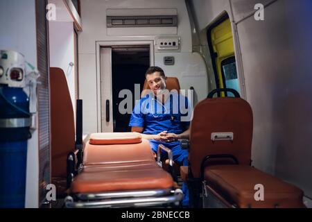 Positivo lavoratore medico maschile seduto in un'auto ambulanza, sorridendo a qualcuno Foto Stock