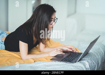 Bella donna brunetta che indossa occhiali e lavora su computer portatile. Donna concentrata che si trova a letto a casa scrivendo e utilizzando la tastiera del notebook Foto Stock