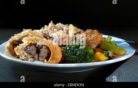 Pollo con broccoli, involtini d'uovo e riso fritto, tradizionale cucina cinese cantonese su un piatto bianco su sfondo nero Foto Stock