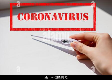 Il francobollo con il segno del coronavirus e il termometro elettronico bianco in mano mostrano basse temperature con luce bianca e sfondo ombreggiato Foto Stock