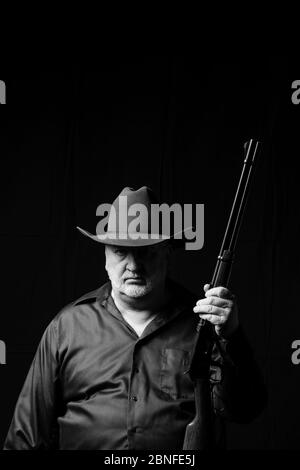 Uomo anziano in un cappello da cowboy con una barba grigia che tiene un fucile a leva con sfondo nero, verticale Foto Stock