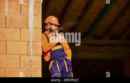 macchina da costruzione fumatrice sigaretta. edificio in costruzione. uomo in uniforme e cappello. copia spazio. ingegnere addetto alla riparazione indossare casco sul posto. costruttore stanco rilassarsi. tempo per rilassarsi. Foto Stock