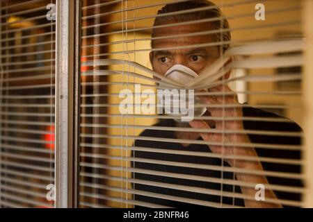 coronavirus, maschio che indossa maschera medica in auto isolamento guardando fuori delle finestre ciechi di casa. L'uomo preoccupato prende le misure di sicurezza di quarantena di Covid 19 Foto Stock