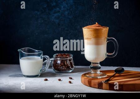 Caffè Dalgona ghiacciato con cacao setacciato su grigio chiaro e sfondo scuro. Caffè alla moda panna montato. Bevanda estiva fredda sudcoreana. Foto Stock