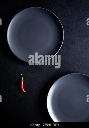 Composizione minimalista di peperoncino rosso e due piatti di ceramica nera al tavolo nero Foto Stock