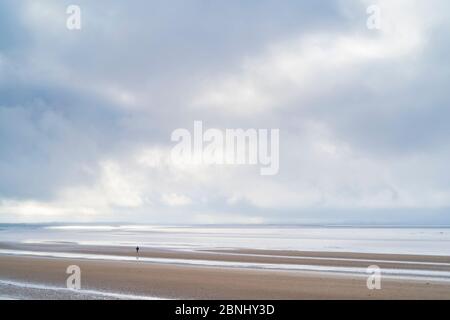 La figura di Lone cammina sulla spiaggia sabbiosa godendo della solitudine lungo la riva del mare vicino al canale di Bristol a Burnham-on-Sea, Somerset, Regno Unito Foto Stock