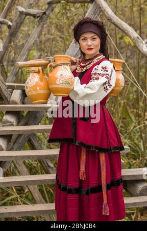 Una donna in abiti tradizionali porta acqua in vecchi barili. Le vecchie caraffe sono trasportate da una ragazza in un vestito stilizzato. Bei vestiti originali del Foto Stock