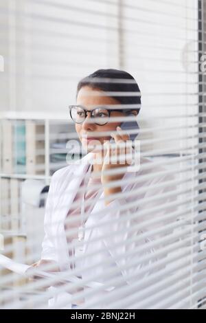 Donna dottore in camice bianco che parla sul telefono cellulare mentre si trova in ufficio dietro l'otturatore Foto Stock