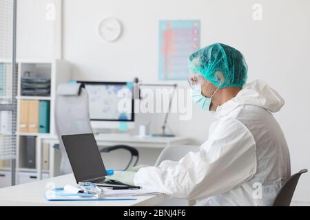 Medico maschile in abbigliamento protettivo seduto sul suo posto di lavoro e digitando sul computer portatile in ufficio Foto Stock