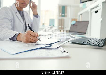 Primo piano di dottore femminile seduto al tavolo e compilando i documenti in ufficio Foto Stock