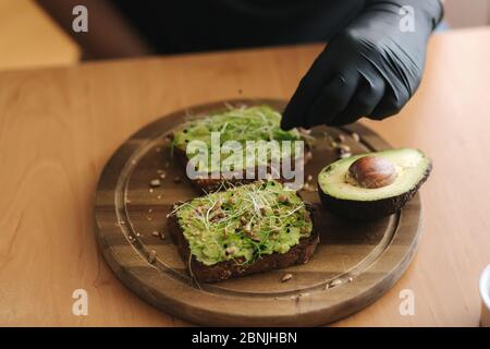 Donna che fa sandwich vegano usando pane tostato di segale con guacamole e micro verdi. Donna cospargere con semi di sesamo su sandwich Foto Stock