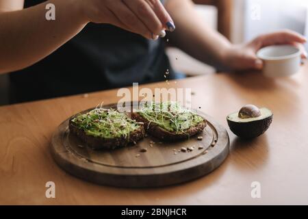 Donna che fa sandwich vegano usando pane tostato di segale con guacamole e micro verdi. Donna cospargere con semi di sesamo su sandwich Foto Stock