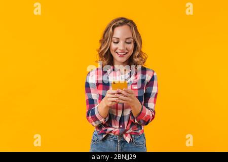 Immagine di giovane bella donna che indossa camicia plaid sorridente e tenendo il cellulare isolato su sfondo giallo Foto Stock