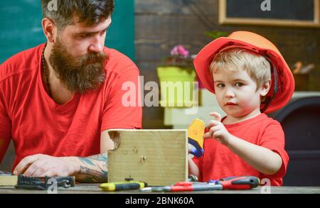 Ragazzo, bambino occupato in casco protettivo imparare ad usare la sega a mano con papà. Padre, genitore con barba insegnando il bambino a segare mentre il figlio gioca con la sega giocattolo. Concetto di giochi educativi Foto Stock