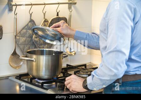 Uomo che cucina in cucina mentre si solleva il coperchio di una pentola sulla stufa mentre il fuoco è acceso. L'uomo indossa una camicia e un jeans blu chiaro. Foto Stock