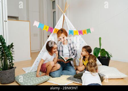 Mamma sorridente impegnata a leggere il letto con i suoi bambini in una capanna a casa Foto Stock