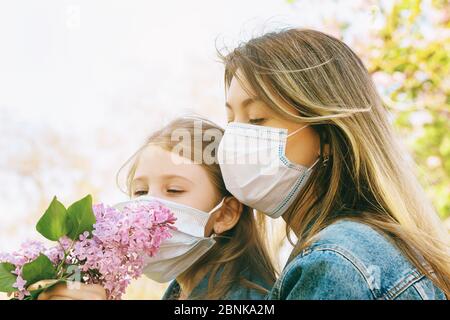 Mamma e figlia in maschere mediche sniff un lilla sulla strada. Concetto di uscita dalla quarantena Foto Stock