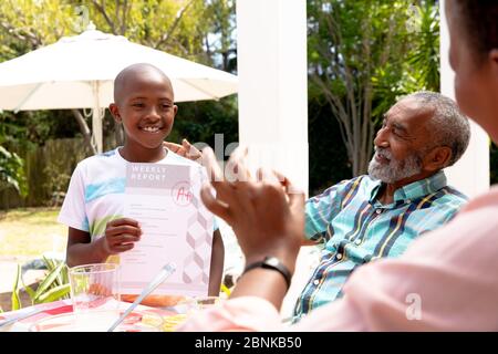 Ragazzo afroamericano che mostra al nonno i suoi compiti durante un pranzo di famiglia in giardino Foto Stock