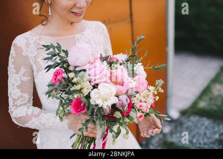 Primo piano di un bouquet di peonie, rose, eucalipto in tonalità bianco-rosa legato a nastri rosa. La sposa tiene un bouquet in mano in un wh Foto Stock