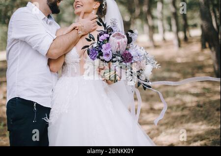 Primo piano del bouquet di rose, garofani, protei e foglie d’argento di una sposa nei toni del bianco, rosa, viola e lavanda. La sposa in un abito bianco pizzo Foto Stock