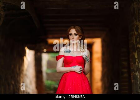 Attraente donna tatuata rossa in lungo vestito rosso e diadema su sfondo sfocato castello medievale Foto Stock