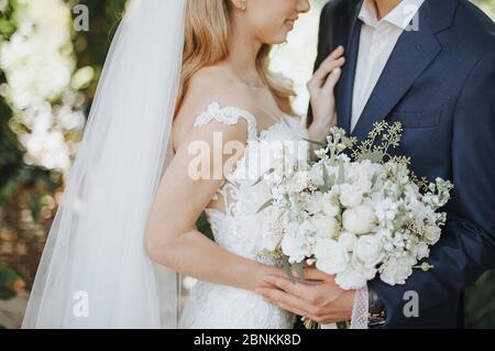 Primo piano del bouquet di fiori bianchi di una sposa. La sposa in un abito bianco pizzo tiene un bouquet in mano. L'abbraccio dello sposo e della sposa, si trova di fronte a ea Foto Stock