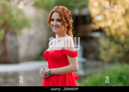 Ritratto di bella donna sorridente tatuata rosso in abito rosso e diadema sulla testa che posa su sfondo moscato castello medievale al sole estivo Foto Stock