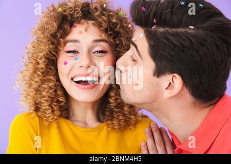 Immagine di simpatico soddisfare coppia sorridente e baciante su guancia mentre ha partito con stelle glitter su volti isolati su sfondo viola Foto Stock