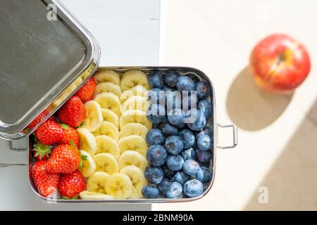 Mirtilli, banane e fragole in un grande contenitore di alimenti in acciaio inossidabile. Scatola pranzo libera in plastica isolata sul tavolo. Mela rossa sullo sfondo. Zero sprechi, ecocompatibile. Foto Stock