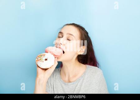 La ragazza giovane tiene due ciambelle in mano e morde uno dal godendo la chiusura dei suoi occhi, sfondo blu Foto Stock